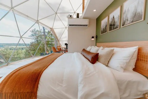 Cozy bedroom in a geodesic dome with a large window, featuring a bed, decorative pillows, and scenic views outside.