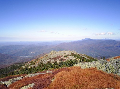 A panoramic view of rolling mountains and valleys, with rocky outcrops and autumn foliage under a clear blue sky.