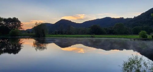 Serene landscape featuring a calm lake reflecting mountains and a colorful sunset, surrounded by lush greenery.