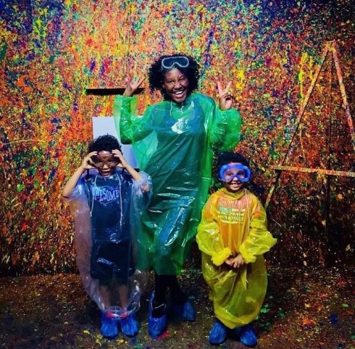 A woman and two children in colorful ponchos and goggles pose playfully in a vibrant, splatter-painted room.
