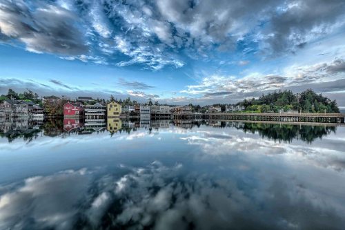 A serene lake reflects colorful houses and a dramatic sky with clouds, creating a peaceful landscape scene.