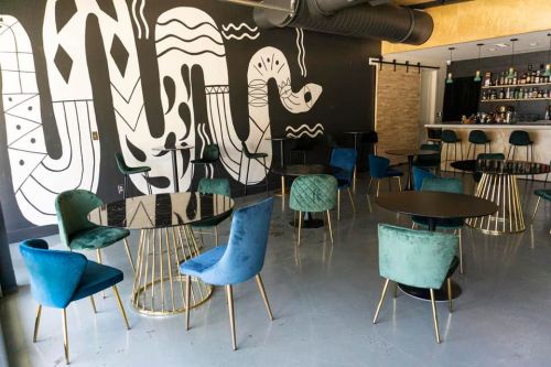 A modern interior featuring round tables with gold legs and colorful chairs, against a black and white mural.