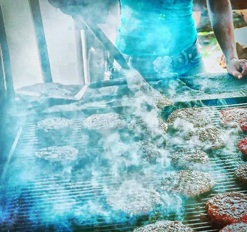 A person grilling burgers on a smoky barbecue, with a spatula in hand and smoke rising from the grill.