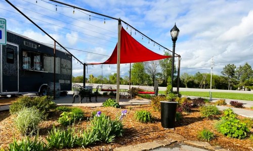 A cozy outdoor seating area with a red canopy, surrounded by flowers and a food truck in the background.