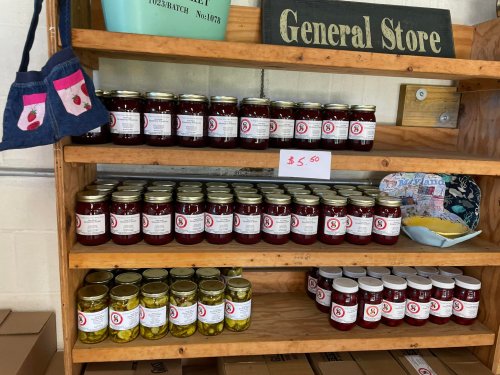 Shelves in a general store display jars of jam and pickles, priced at $5.60, with a blue bag hanging nearby.
