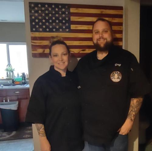 A man and woman in chef uniforms stand together in a kitchen, with an American flag art piece on the wall behind them.