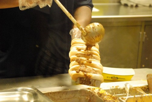A person in gloves serves chili over a row of hot dogs in a kitchen setting.