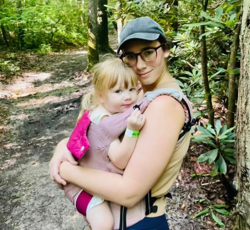A woman holds a young girl in a carrier while walking on a forest trail, surrounded by greenery.