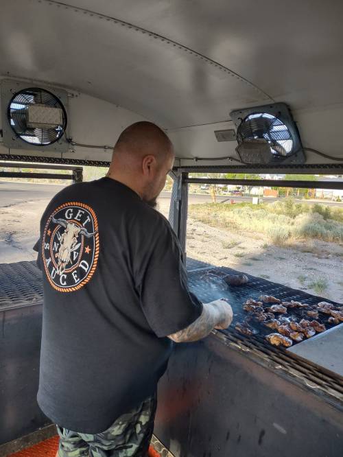 A person with a tattooed arm cooks meat on a grill inside a food truck, wearing a black shirt with a logo.