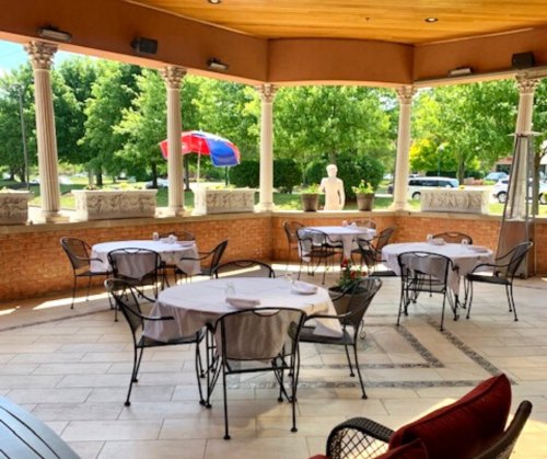 Outdoor dining area with empty tables, surrounded by greenery and decorative columns, under a wooden ceiling.