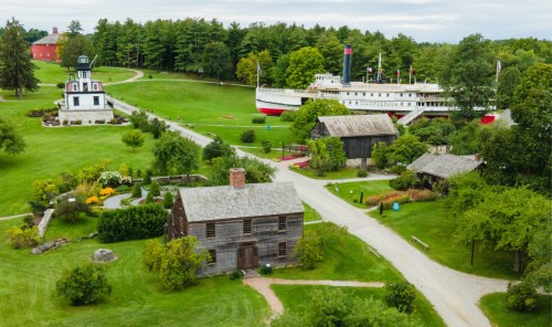 Aerial view of a historic village with a wooden house, gardens, and a large ship docked nearby, surrounded by greenery.