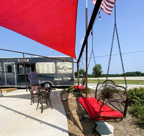 A food truck with a menu, shaded seating area, and a person standing nearby under a red canopy.