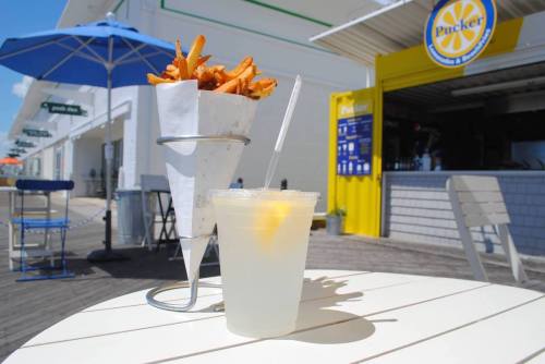 A cup of lemonade with a slice of lemon beside a cone of sweet potato fries on a table outdoors.