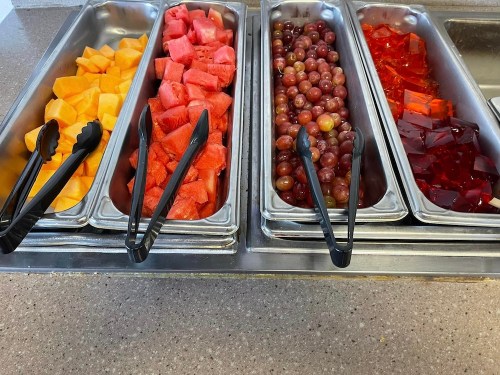 A buffet display with trays of cantaloupe, watermelon, grapes, and red gelatin dessert. Tongs are placed in each tray.