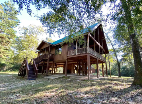 A wooden house on stilts surrounded by trees, featuring a blue roof and a staircase leading to the entrance.