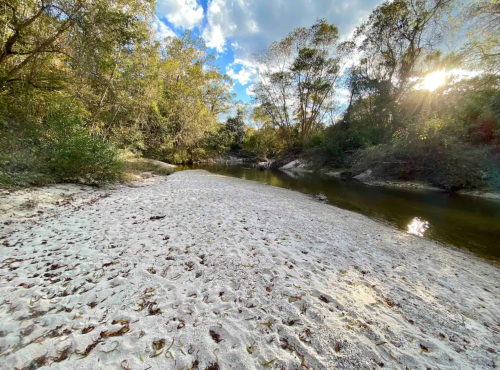 A serene riverbank scene with sandy shores, surrounded by trees and sunlight filtering through the clouds.