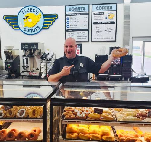 A smiling man in a uniform holds a donut, standing behind a display case at Flyboy Donuts. Donut menu visible in the background.