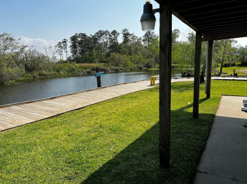 A peaceful view of a riverbank with a wooden dock, surrounded by greenery and a clear blue sky.