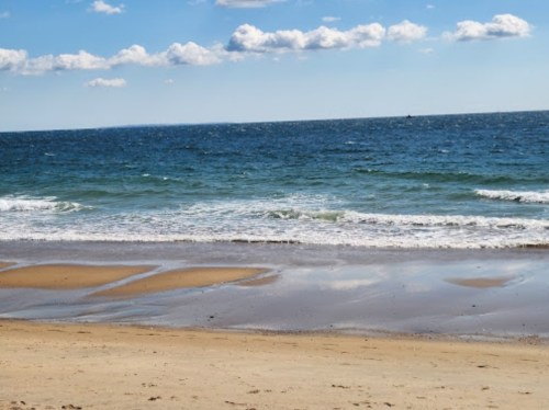 A serene beach scene with gentle waves lapping at the shore under a blue sky with scattered clouds.