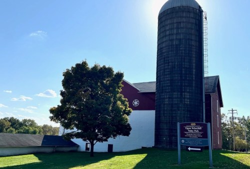 A red barn with a silo, surrounded by green grass and a tree, under a clear blue sky. A sign points to a nearby park.