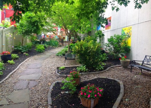A serene garden path lined with flowers, greenery, and stone, leading to a cozy seating area.