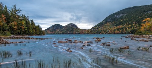 A serene lake surrounded by mountains and colorful autumn foliage under a cloudy sky. Rocks and grass are visible in the water.