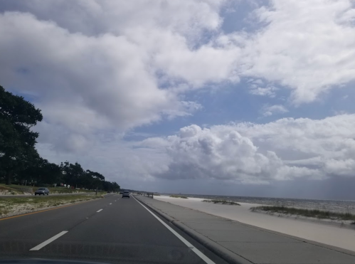 A scenic view of a road alongside a beach, with clouds in the sky and sand dunes visible on the shore.