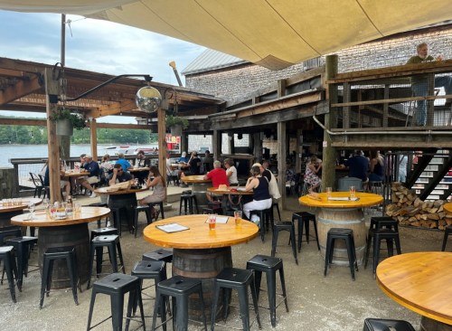 Outdoor dining area with wooden tables and stools, people enjoying meals, and a view of the water in the background.