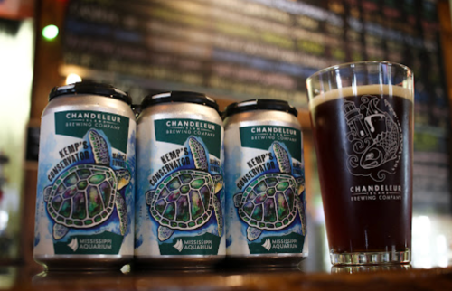 Three cans of "Kemp's Conservation" beer next to a glass with the Chandeleur Brewing Company logo on a bar.