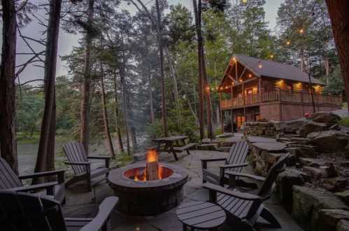 Cozy cabin in the woods with a fire pit and seating area, surrounded by trees and a river in the background.