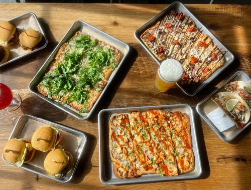 A spread of various pizzas, sliders, and a drink on metal trays atop a wooden table.
