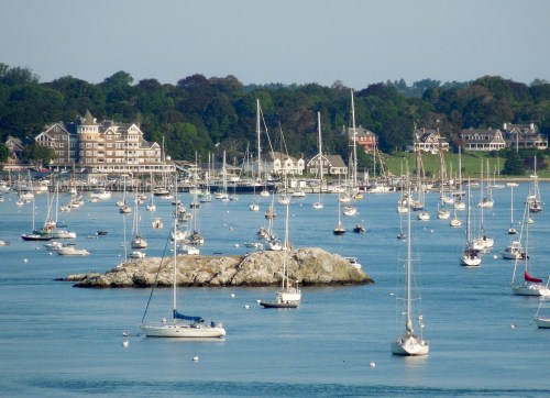 A serene harbor filled with sailboats, rocky islets, and lush green hills in the background under a clear blue sky.