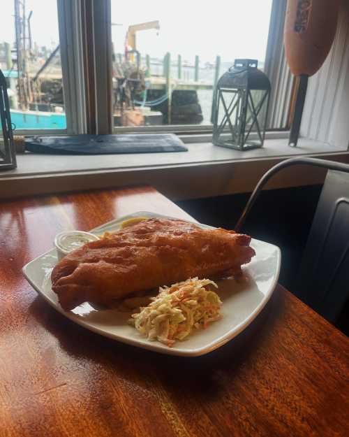 A plate of fried fish with coleslaw and dipping sauce, set by a window overlooking a harbor.