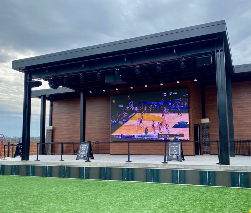 A large outdoor screen displays a basketball game on a modern stage with a grassy area in front.