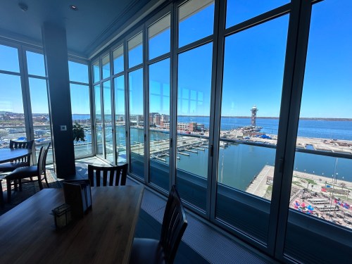 A bright, modern dining area with large windows overlooking a marina and waterfront under a clear blue sky.