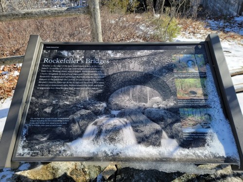 A sign about Rockefeller's Bridges, featuring text and images, surrounded by snow-covered rocks and trees.