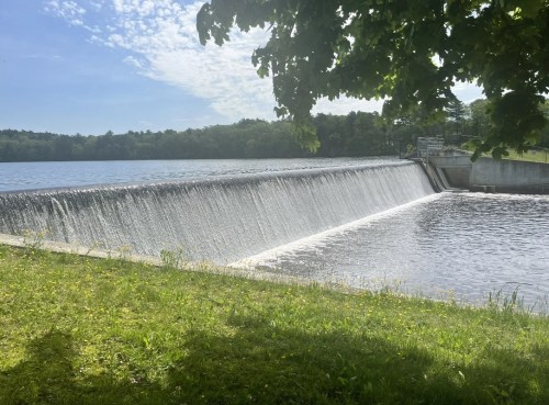 A serene view of a dam with water cascading over the edge into a calm lake, surrounded by greenery and blue skies.