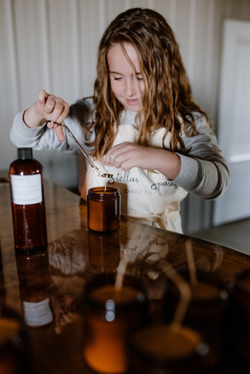 A young girl in an apron carefully adds a wick to a candle jar while surrounded by other candle-making supplies.
