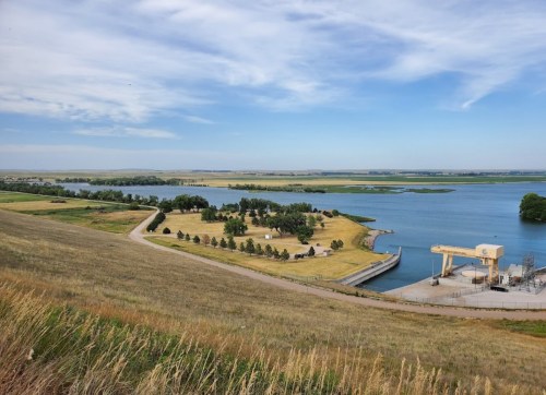 A scenic view of a reservoir with green fields, trees, and a winding road under a blue sky with scattered clouds.
