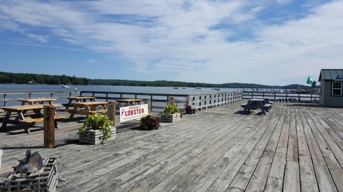 A wooden pier with picnic tables, lobster sign, and a view of the water and boats under a blue sky.