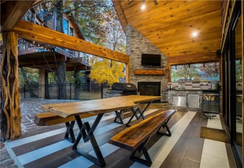 Outdoor dining area with a wooden table, benches, stone fireplace, and grill, surrounded by trees and autumn foliage.