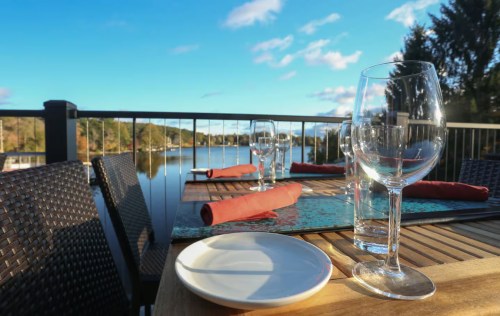 A scenic outdoor dining setup with glasses and a plate, overlooking a calm lake and trees under a blue sky.
