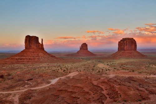 Three iconic sandstone buttes rise from the desert landscape under a colorful sunset sky.
