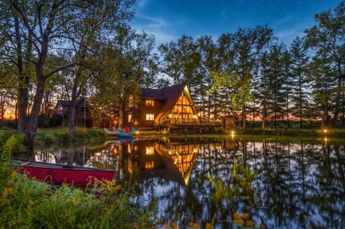 A serene lakeside scene featuring a cozy A-frame cabin reflecting in the water at dusk, surrounded by trees.