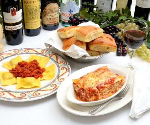 A plate of ravioli with sauce, a serving of lasagna, bread rolls, and a glass of wine, surrounded by wine bottles and grapes.