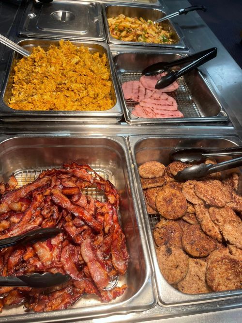 A buffet display featuring scrambled eggs, sliced ham, crispy bacon, and sausage patties in metal trays.