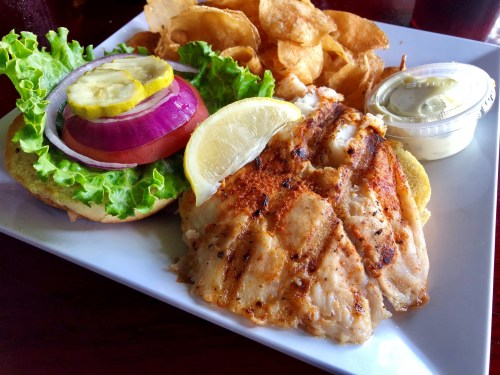 A plate featuring grilled fish with lemon, a sandwich with lettuce, tomato, onion, pickles, and a side of potato chips.