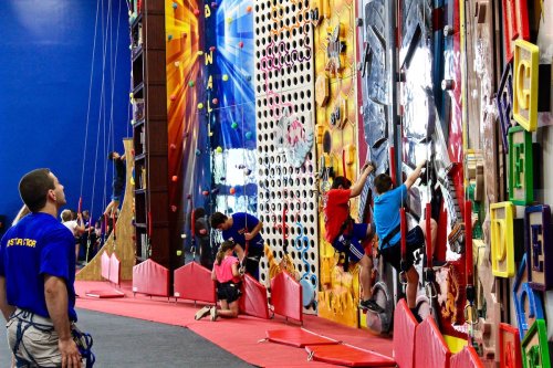 A climbing gym with people scaling various walls, while others observe and assist from the ground.