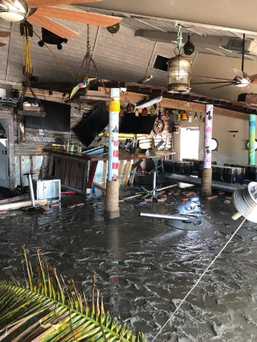 Interior of a flooded bar with debris, overturned furniture, and water covering the floor, creating a chaotic scene.