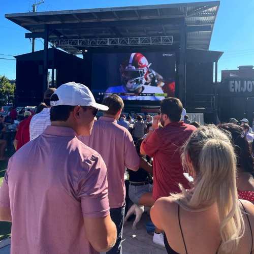 A crowd watches a large outdoor screen displaying a football game, with people in casual attire enjoying the event.